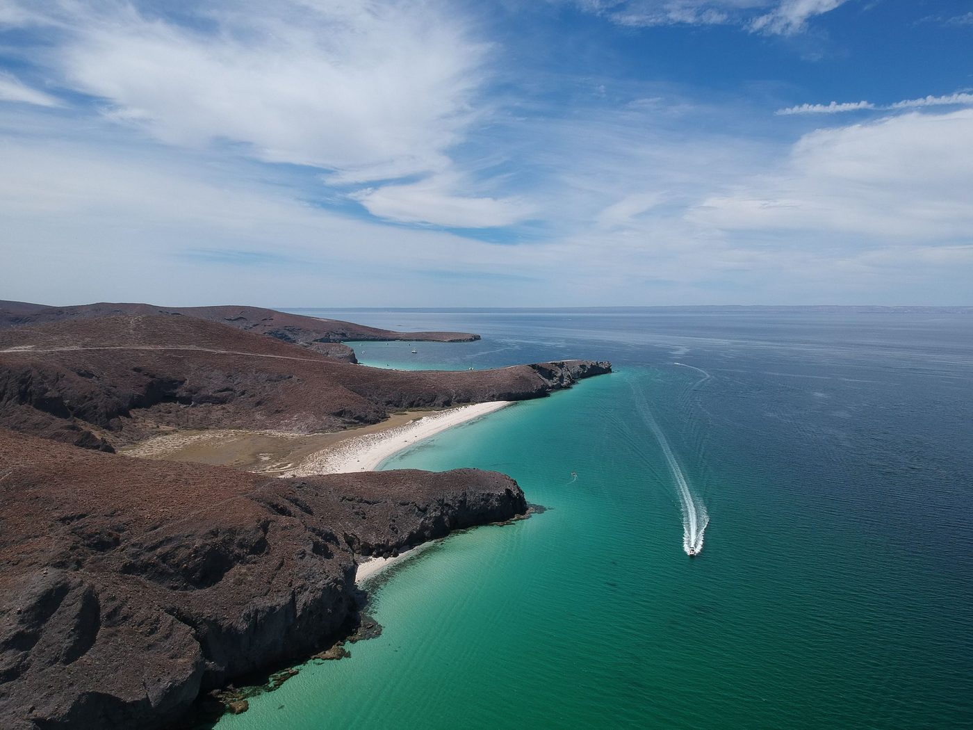 Aerial view of dark desert cliffs and a white beach above turquoise water, a boat wake offshore
