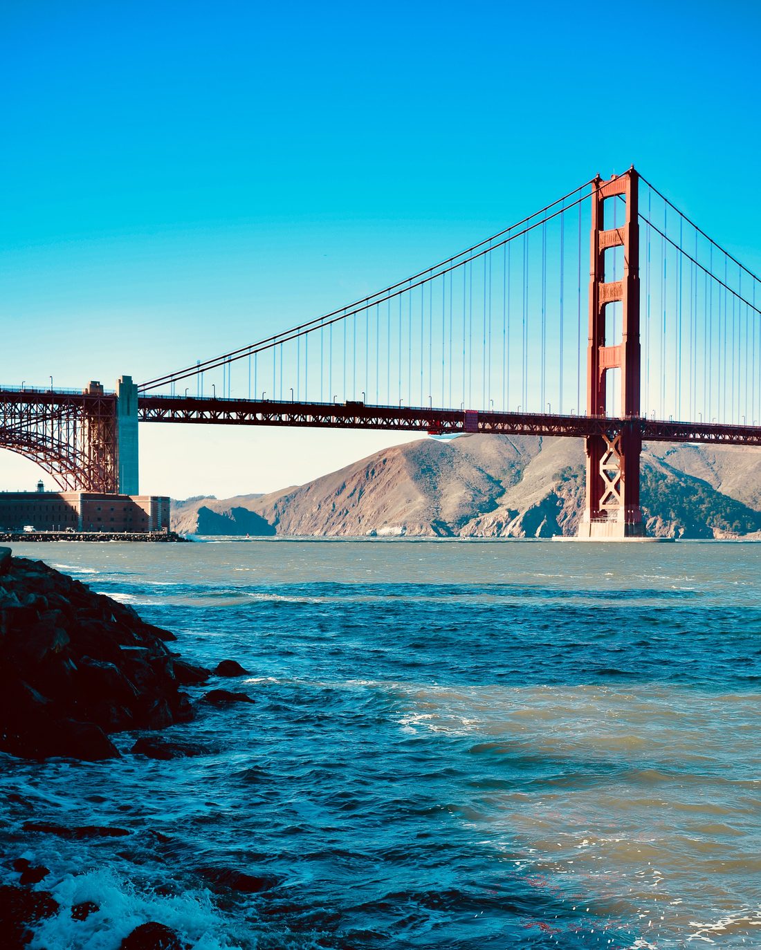 The Golden Gate Bridge over the harbor entrance, rocks and surf in the foreground