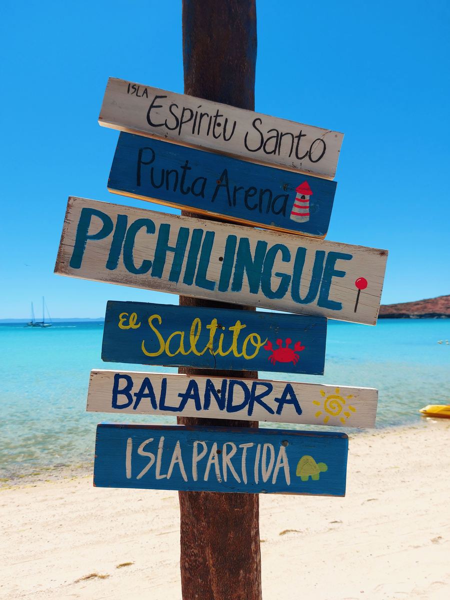 Hand-painted signpost on the beach listing Espíritu Santo, Pichilingue, Balandra and Isla Partida